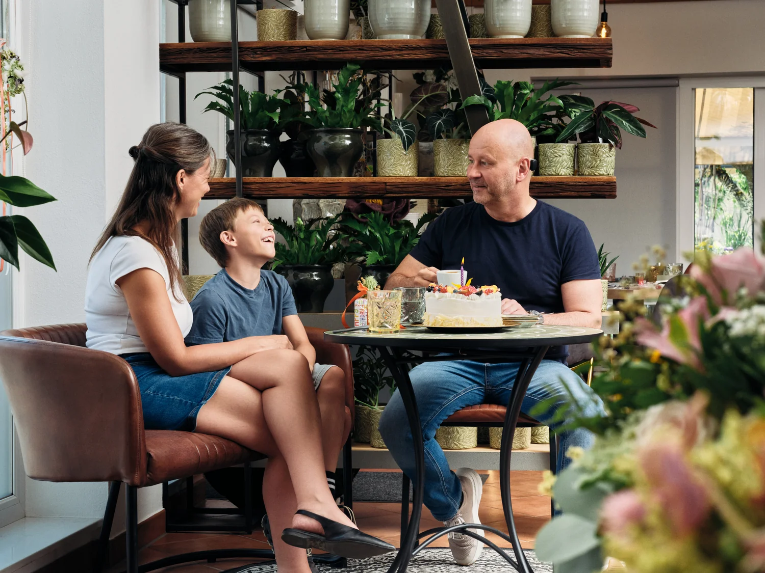 Vater, Mutter und Kind sitzen im Kaffeehaus, Geburtstagstorte steht auf dem Tisch. Vater trägt Hörgerät.